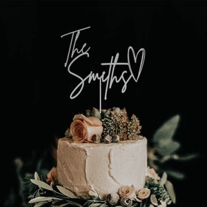 May include: A white wedding cake with a silver cake topper that says "The Smiths" with a heart. The cake is decorated with greenery and a single peach-coloured rose.