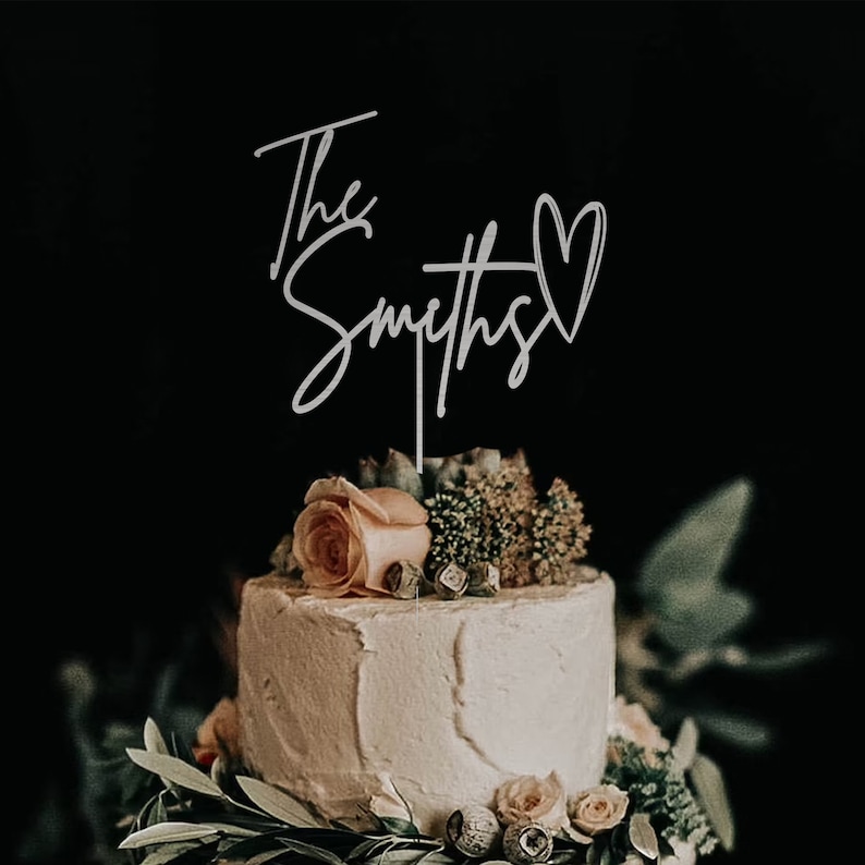May include: A white wedding cake with a silver cake topper that says "The Smiths" with a heart. The cake is decorated with greenery and a single peach-coloured rose.