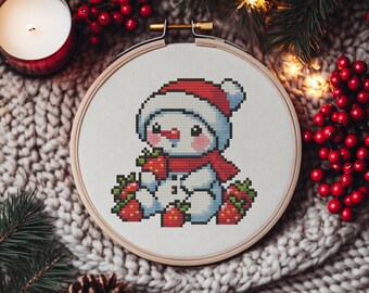 May include: A cross-stitch embroidery hoop featuring a cute snowman wearing a red hat and scarf, surrounded by strawberries. The snowman is stitched on white fabric, set in a wooden hoop. The background includes pine needles, red berries, and a lit candle.