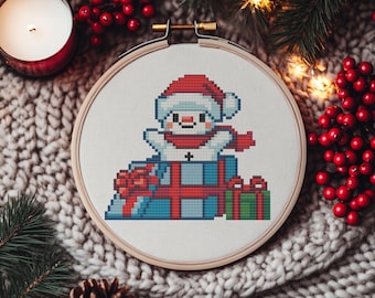 May include: A cross-stitch embroidery hoop featuring a pixelated snowman wearing a Santa hat and scarf, surrounded by wrapped gifts. The snowman is atop a pile of blue and red wrapped presents. The embroidery is set against a cozy, textured background.