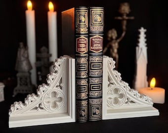 May include: Two antique books with black covers and gold lettering are held by a pair of white gothic-style bookends. The bookends feature an ornate design. Lit candles and other decorative items are in the background, creating a dark, atmospheric scene.