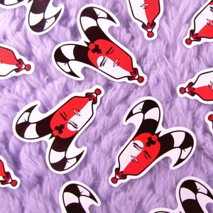 Puede incluir: Una colección de pegatinas con una cara estilizada con una combinación de colores rojo y blanco, una cruz negra y un sombrero de bufón con rayas blancas y negras. Las pegatinas están esparcidas sobre una superficie púrpura suave.