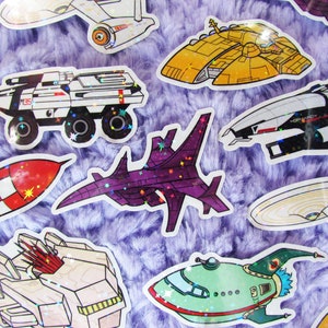 Könnte beinhalten: Eine Sammlung farbenfroher, holografischer Aufkleber mit verschiedenen Raumschiffdesigns. Die Aufkleber zeigen ein weißes und schwarzes gepanzertes Fahrzeug, eine rot-weiße Rakete und mehrere futuristische Raumschiffe in Gelb-, Lila- und Grüntönen.