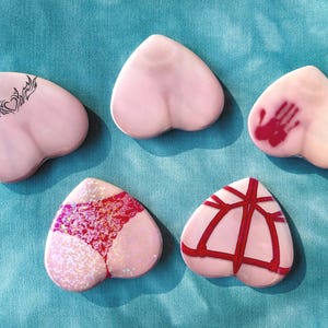 Könnte beinhalten: Fünf herzförmige Objekte in Rosa mit verschiedenen Designs. Eines hat ein schwarzes Stammes-Tattoo, ein anderes einen roten Handabdruck, und ein drittes zeigt rote Glitzerspitze. Die restlichen zwei haben kein Design und rote Linien.