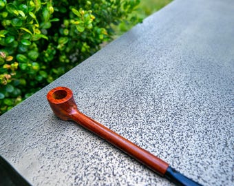May include: A wooden pipe with a dark brown stem and a reddish-brown bowl. The pipe rests on a textured, gray surface, with green foliage in the background. The pipe's mouthpiece is black.