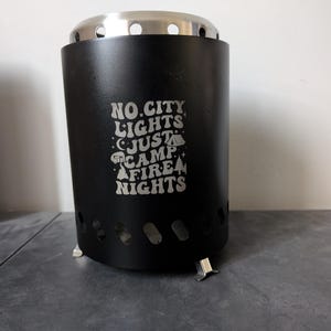 May include: Black metal camping stove with a silver top and three legs. The stove has a graphic that reads "No City Lights Just Camp Fire Nights".