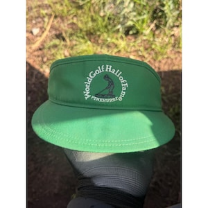 May include: A green visor with the text "World Golf Hall of Fame" in white lettering. The visor has a curved brim and a circular logo featuring a golfer. The text "PINEHURST" is also visible.