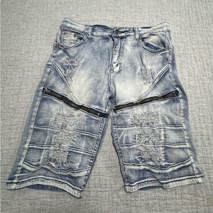 May include: Light blue denim shorts with distressed detailing and black zippers. The shorts feature a classic five-pocket design and a button and zipper closure. The denim has a faded wash and a worn appearance.