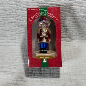 May include: A Christmas Treasures hand-crafted glass ornament of a nutcracker soldier in a red box. The nutcracker is wearing a red and gold jacket, blue pants, and a tricorn hat. The box has the words "Christmas Treasures" and "Classic Angels".