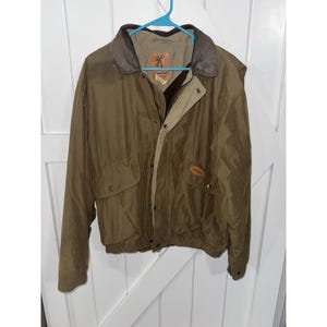 May include: Olive green jacket with a brown collar and tan lining. The jacket has two front pockets, a button closure, and a small logo on the chest. The jacket is hanging on a blue hanger.