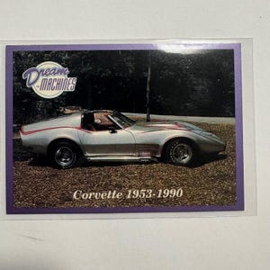 Peut inclure: Carte de collection représentant une Corvette argentée décapotable avec des détails rouges. La carte porte les inscriptions "Dream Machines" et "Corvette 1953-1990". La voiture est garée sur des cailloux bruns.