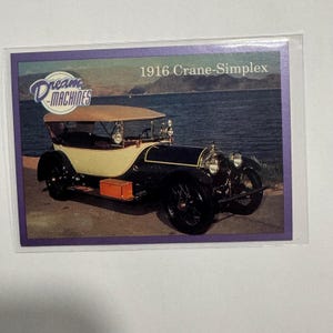 May include: A trading card depicting a 1916 Crane-Simplex car. The classic car is black and cream with a tan convertible roof. The card features the text "Dream Machines" and "1916 Crane-Simplex". The background shows a body of water and a shoreline.