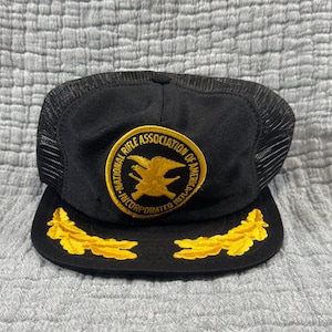 May include: Black trucker hat with a mesh back and a curved brim. The front features a gold emblem with the text "NATIONAL RIFLE ASSOCIATION OF AMERICA INCORPORATED 1871". Gold leaf designs adorn the brim.