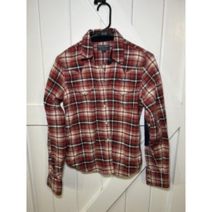 Pode incluir: Uma camisa xadrez vermelha, preta e branca com mangas compridas. A camisa tem uma gola clássica, dois bolsos no peito e um pequeno patch na parte superior do peito. A camisa está pendurada em um cabide preto.
