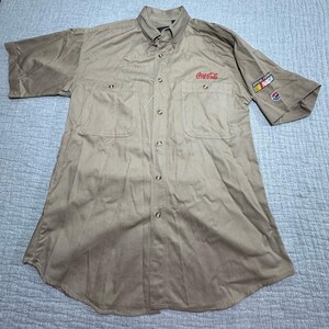 May include: Khaki short-sleeved button-down shirt with two chest pockets. Features the Coca-Cola logo in red on the right chest and a NASCAR patch on the left sleeve. The shirt has a button-down collar.