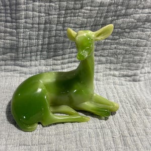 May include: A green glass deer figurine in a resting pose. The deer has a glossy finish and is a vibrant shade of green, with lighter green accents on the ears and legs. The figurine is a decorative item.