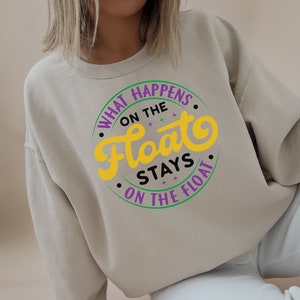 May include: Beige sweatshirt with a circular graphic that reads "WHAT HAPPENS ON THE FLOAT STAYS ON THE FLOAT" in purple, green, yellow, and black. The sweatshirt is a neutral color and has long sleeves.