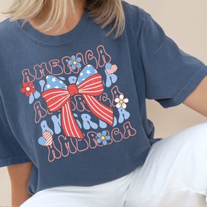 Pode incluir: Uma camiseta azul marinho com um laço vermelho, branco e azul e o texto "America" em uma fonte retrô.