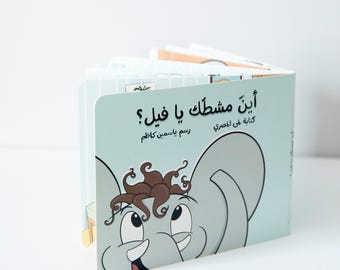 May include: A light blue children's book with the title "Aish Mishtak Ya Fil?" in Arabic script. The book features a cartoon illustration of an elephant with brown curly hair and a big smile.