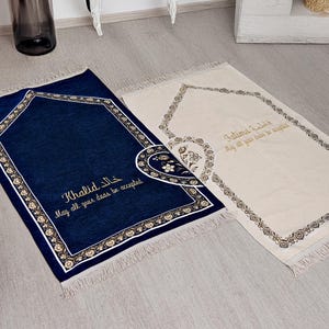 Puede incluir: Se muestran dos alfombras de oración, una azul marino y otra crema. La alfombra azul marino presenta ribetes dorados y el nombre "Khalid" en escritura dorada. La alfombra crema tiene el nombre "Fatima" en escritura dorada. Ambas tienen bordes decorativos y flecos.