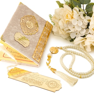 May include: A beige velvet-covered Quran with gold accents and a personalized nameplate. The Quran is open to reveal Arabic text. A matching bookmark and a white pearl prayer beads with a gold tassel are also included.