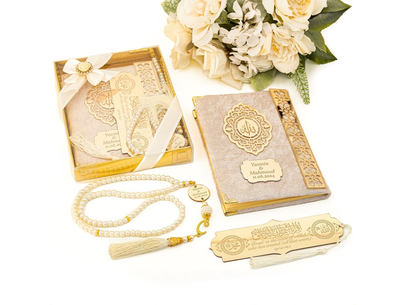 May include: A gold-trimmed gift box containing a beige velvet prayer book with a gold-colored inscription that reads "Yasmin & Mahmoud 31.08.2024". The box also includes a gold-colored bookmark with Arabic calligraphy and a white pearl prayer beads with a tassel.