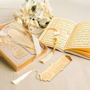 May include: A gold gift box with a white ribbon and a white tassel. The box contains a gold-colored bookmark with a tassel and a gold-colored prayer beads. The box also has a personalized name tag with the text "Yasmin & Mahmoud 1.06.2004".