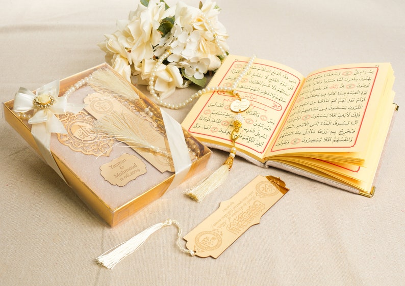 May include: A gold gift box with a white ribbon and a white tassel. The box contains a gold-colored bookmark with a tassel and a gold-colored prayer beads. The box also has a personalized name tag with the text "Yasmin & Mahmoud 1.06.2004".