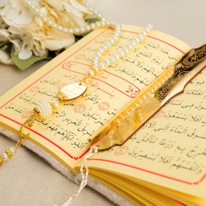 May include: An open Quran with a gold bookmark and a white pearl prayer beads with a gold tassel. The book is on a white surface.