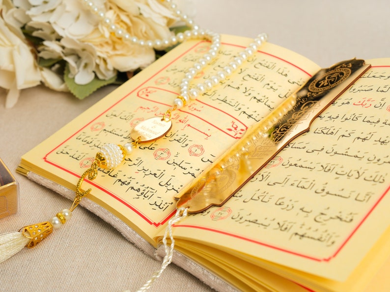 May include: An open Quran with a gold bookmark and a white pearl prayer beads with a gold tassel. The book is on a white surface.