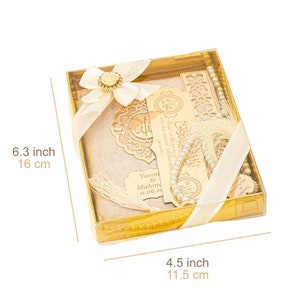May include: A gold gift box with a white satin ribbon and a white prayer rug inside. The box also contains a wooden bookmark with Arabic calligraphy, a set of prayer beads, and a personalized wooden tag. The box measures 4.5 inches by 6.3 inches.