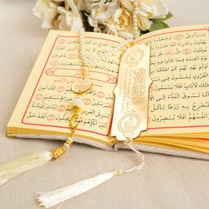 May include: A gold-colored bookmark with Arabic calligraphy and a tassel hangs from a white and gold prayer bead necklace. The necklace is resting on an open book with Arabic text.