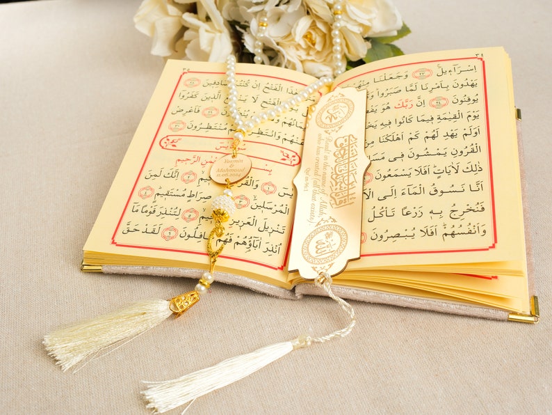 May include: A gold-colored bookmark with Arabic calligraphy and a tassel hangs from a white and gold prayer bead necklace. The necklace is resting on an open book with Arabic text.