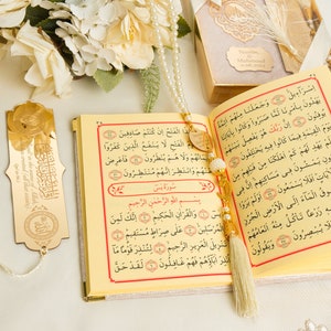 May include: A gold-colored bookmark with Arabic calligraphy and a white tassel hangs from a gold-colored Quran with white pearl beads and a white tassel. The Quran is open to a page with Arabic text.