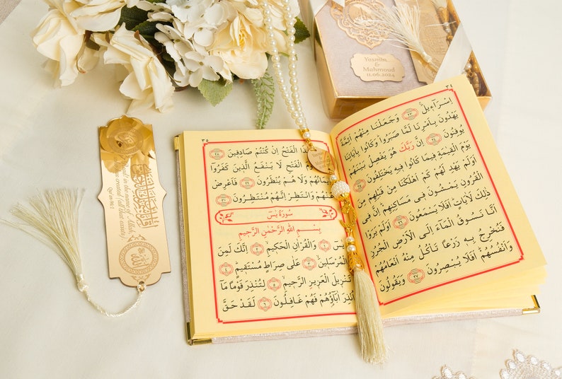 May include: A gold-colored bookmark with Arabic calligraphy and a white tassel hangs from a gold-colored Quran with white pearl beads and a white tassel. The Quran is open to a page with Arabic text.