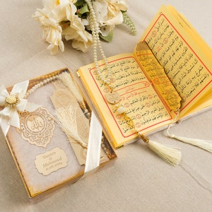 May include: A white gift box with a gold trim and a white ribbon. Inside the box is a white prayer rug, a gold bookmark with a tassel, and a white prayer beads. The box also has a gold tag with the text "Yasmin & Mahmoud 11.06.2024".