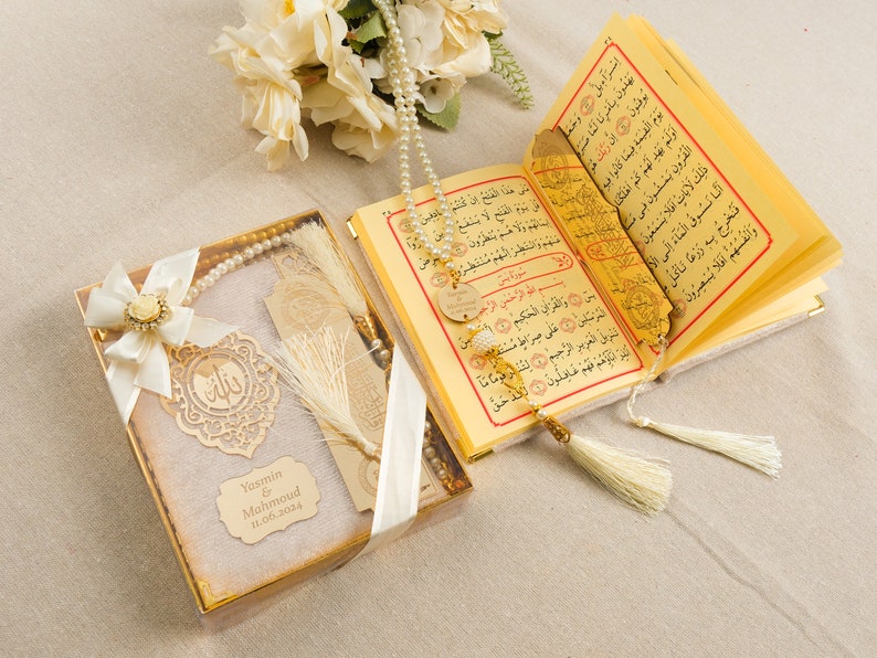 May include: A white gift box with a gold trim and a white ribbon. Inside the box is a white prayer rug, a gold bookmark with a tassel, and a white prayer beads. The box also has a gold tag with the text "Yasmin & Mahmoud 11.06.2024".