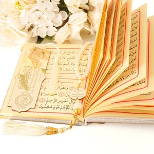 May include: An open book with a white tassel bookmark and a beaded chain with a gold charm that says "Yamin Muhammad". The book has Arabic text on the pages and a gold-colored bookmark with Arabic text.