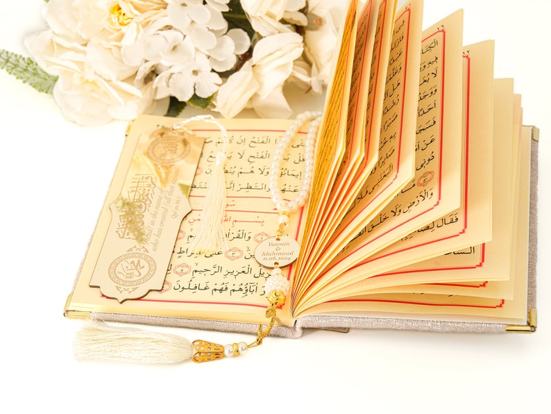 May include: An open book with a white tassel bookmark and a beaded chain with a gold charm that says "Yamin Muhammad". The book has Arabic text on the pages and a gold-colored bookmark with Arabic text.