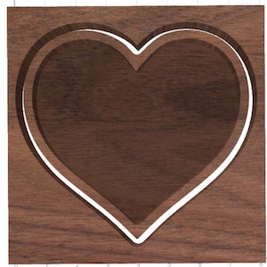 Può includere: Una targa in legno con una forma di cuore a doppio strato scolpita. Il cuore è contornato di bianco e il legno è di colore marrone scuro.