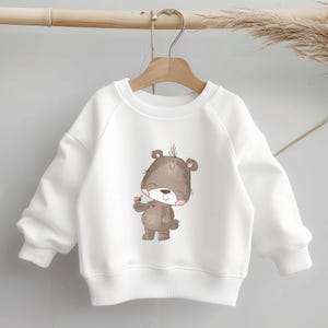 Könnte beinhalten: Weißer Pullover mit einem braunen Bären, der eine rosa Blume hält.