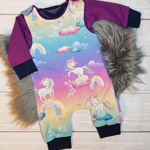 Könnte beinhalten: Ein Baby-Strampler mit einem Regenbogen-Farbverlauf und einem Einhorn-Muster. Der Strampler hat lange Ärmel und einen dunkelblauen Saum. Der Strampler ist aus weichem, bequemem Stoff.