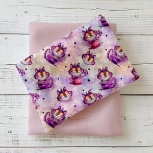 Könnte beinhalten: Zwei Stoffstücke. Der obere Stoff hat ein sich wiederholendes Muster aus lila Cheshire Cat-Illustrationen mit einem breiten Grinsen, vor einem pastellfarbenen Hintergrund. Der untere Stoff ist einfarbig hellrosa. Die Stoffe liegen auf einer weißen Holzoberfläche.