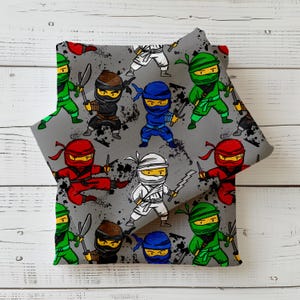 Könnte beinhalten: Grauer Stoff mit einem sich wiederholenden Muster von Cartoon-Ninjas in verschiedenen Farben, darunter Rot, Grün, Blau, Weiß und Braun. Jeder Ninja hält ein Schwert. Der Stoff ist gefaltet und zeigt das Muster auf beiden Seiten.