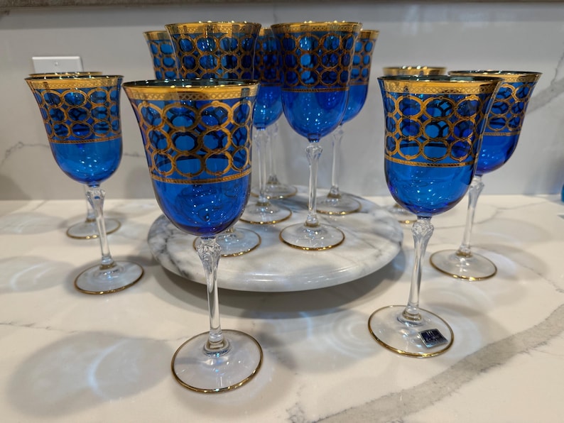 May include: A collection of blue wine glasses with gold rims and intricate gold geometric patterns. The glasses are arranged on a white marble serving tray, showcasing their elegant design. The stemware is ideal for special occasions.