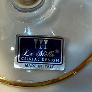 May include: Close-up of a clear glass with a gold rim. A label on the glass reads "Le Si&egrave;lle Cristal Design Made in Italy". The glass has a stem and a wide bowl.
