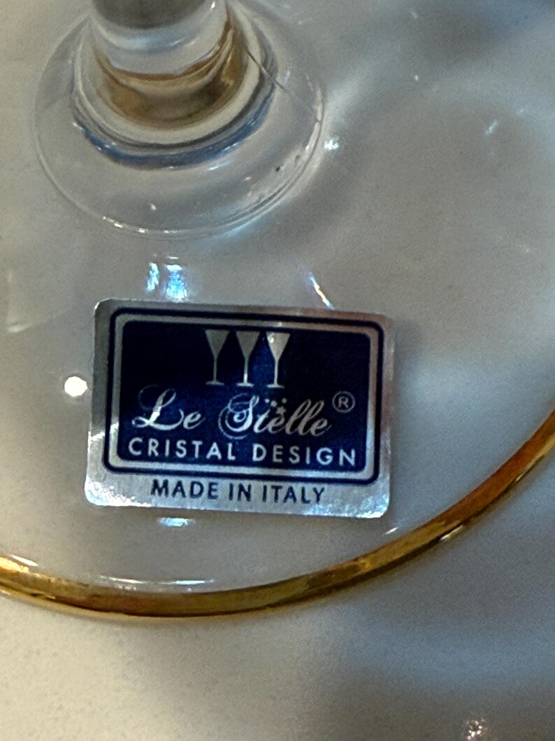 May include: Close-up of a clear glass with a gold rim. A label on the glass reads "Le Si&egrave;lle Cristal Design Made in Italy". The glass has a stem and a wide bowl.