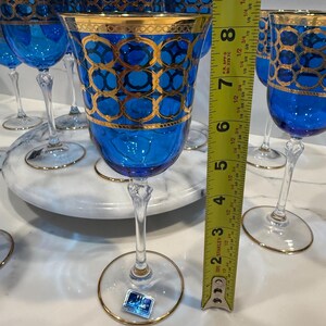 May include: A collection of blue crystal wine glasses with gold trim and decorative gold patterns. The glasses are approximately 7 inches tall. The stemware is displayed on a marble surface.