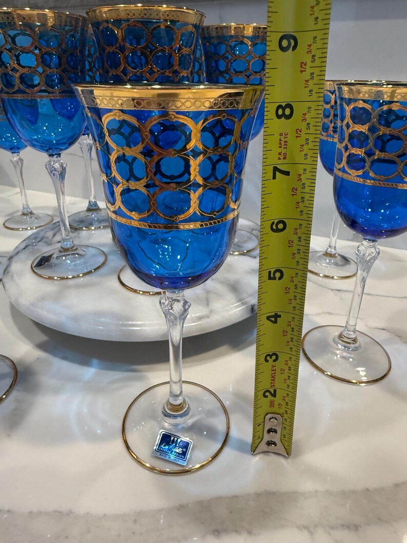May include: A collection of blue crystal wine glasses with gold trim and decorative gold patterns. The glasses are approximately 7 inches tall. The stemware is displayed on a marble surface.