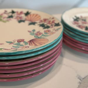 May include: A set of ceramic plates with a cream-colored surface, featuring a seashell and floral design. The plates have a pink rim and a teal rim. The plates are stacked in two separate piles.